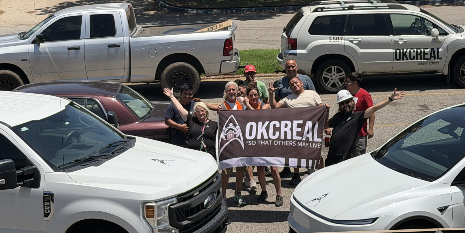 The OKCREAL crew and office team on a campus day, holding the OKCREAL flag between the service trucks